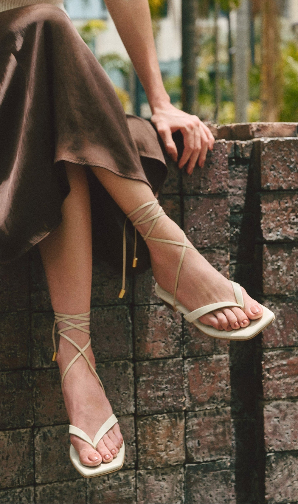 Person wearing a brown skirt and white cream leather sandals sitting on a stone wall.