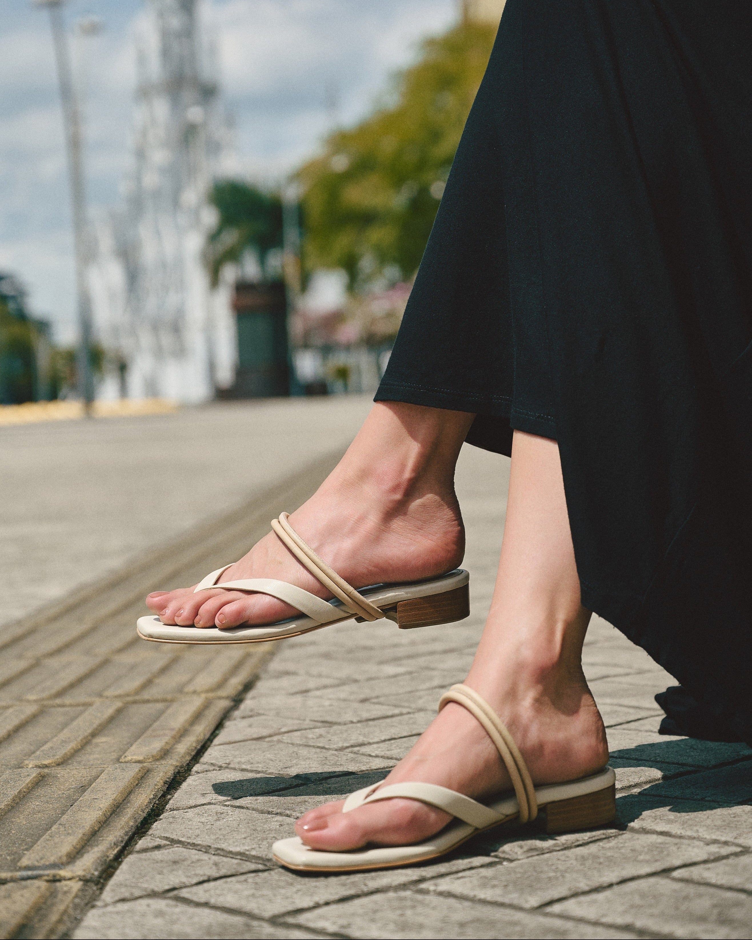 Person wearing beige leather sandals on a sidewalk with a blurred urban background